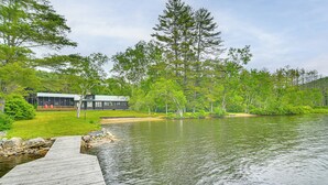 A wooden dock leading to a house with a green roof, surrounded by lush greenery and a calm lake.
