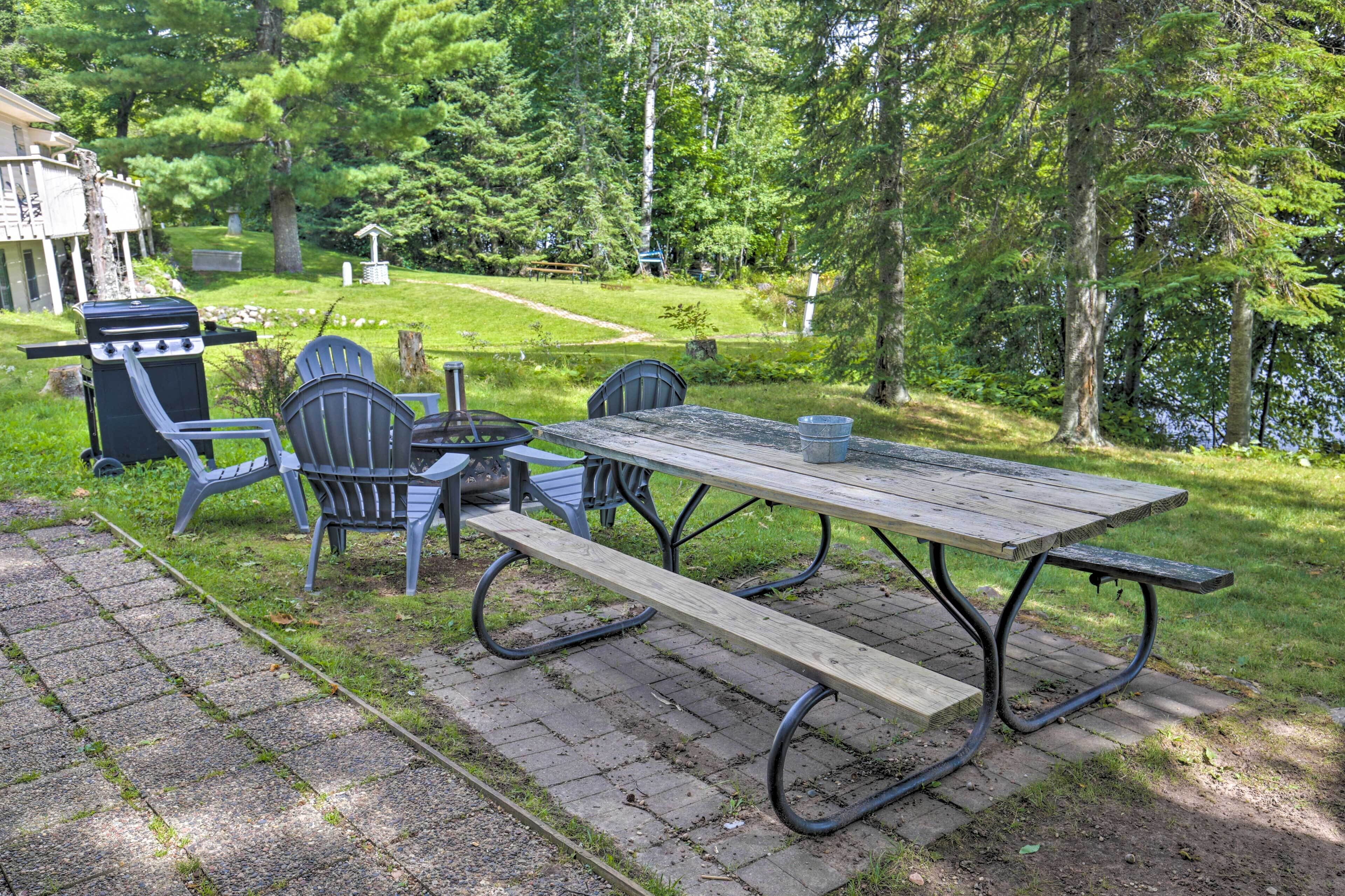 A picnic table with chairs and a grill in a grassy area with trees and a building in the background.