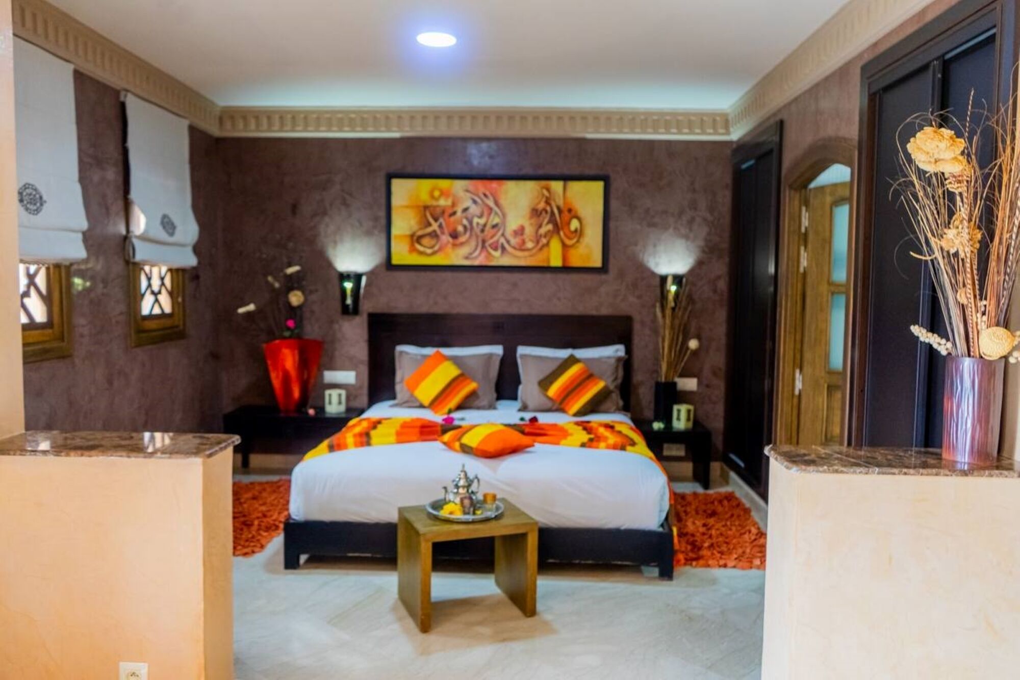 A hotel room with a large bed, a bedside table, a nightstand, a decorative wall painting, and a vase with dried plants.