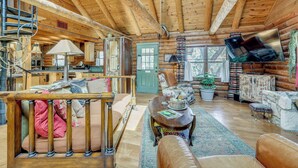 Een blokhut interieur met een houten trap, een eethoek, een woonkamer met een bank en fauteuils, en een aan de muur bevestigde televisie.