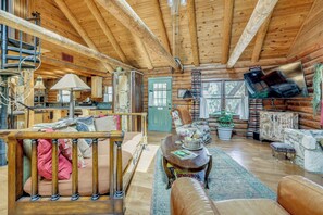 Ein Interieur einer Blockhütte mit einer Holztreppe, einem Essbereich, einem Wohnzimmer mit Sofa und Sesseln sowie einem an der Wand montierten Fernseher.