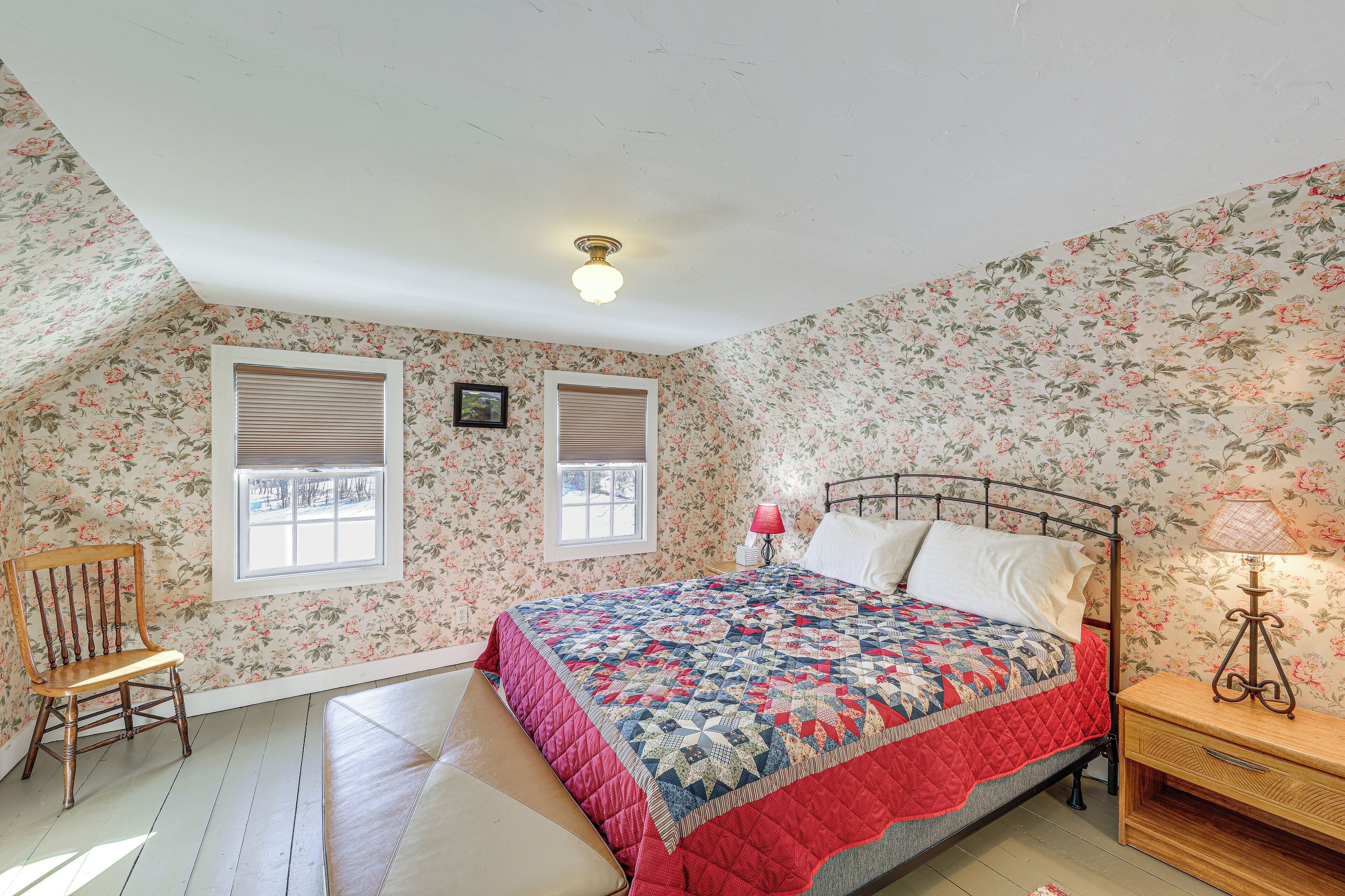 A bedroom with a bed, two windows with blinds, a chair, a bedside table with a lamp, and floral wallpaper.