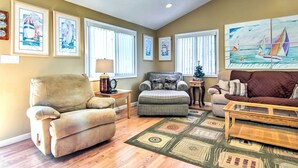 A living room with a beige armchair, a gray sofa, and a brown sofa. There are framed pictures on the wall and a large painting of sailboats.