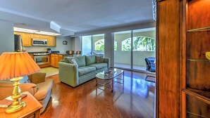 A spacious living room with a sofa, coffee table, and lamp. A kitchen is visible in the background with cabinets and appliances. - Marco Island Condo w/ Patio: Steps to Beach Access (Marco Island)
