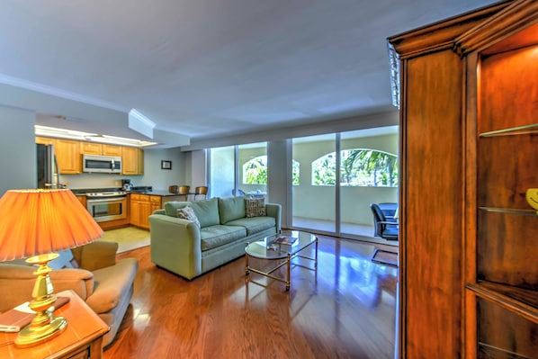 A spacious living room with a sofa, coffee table, and lamp. A kitchen is visible in the background with cabinets and appliances. - Marco Island Condo w/ Patio: Steps to Beach Access (Marco Island)