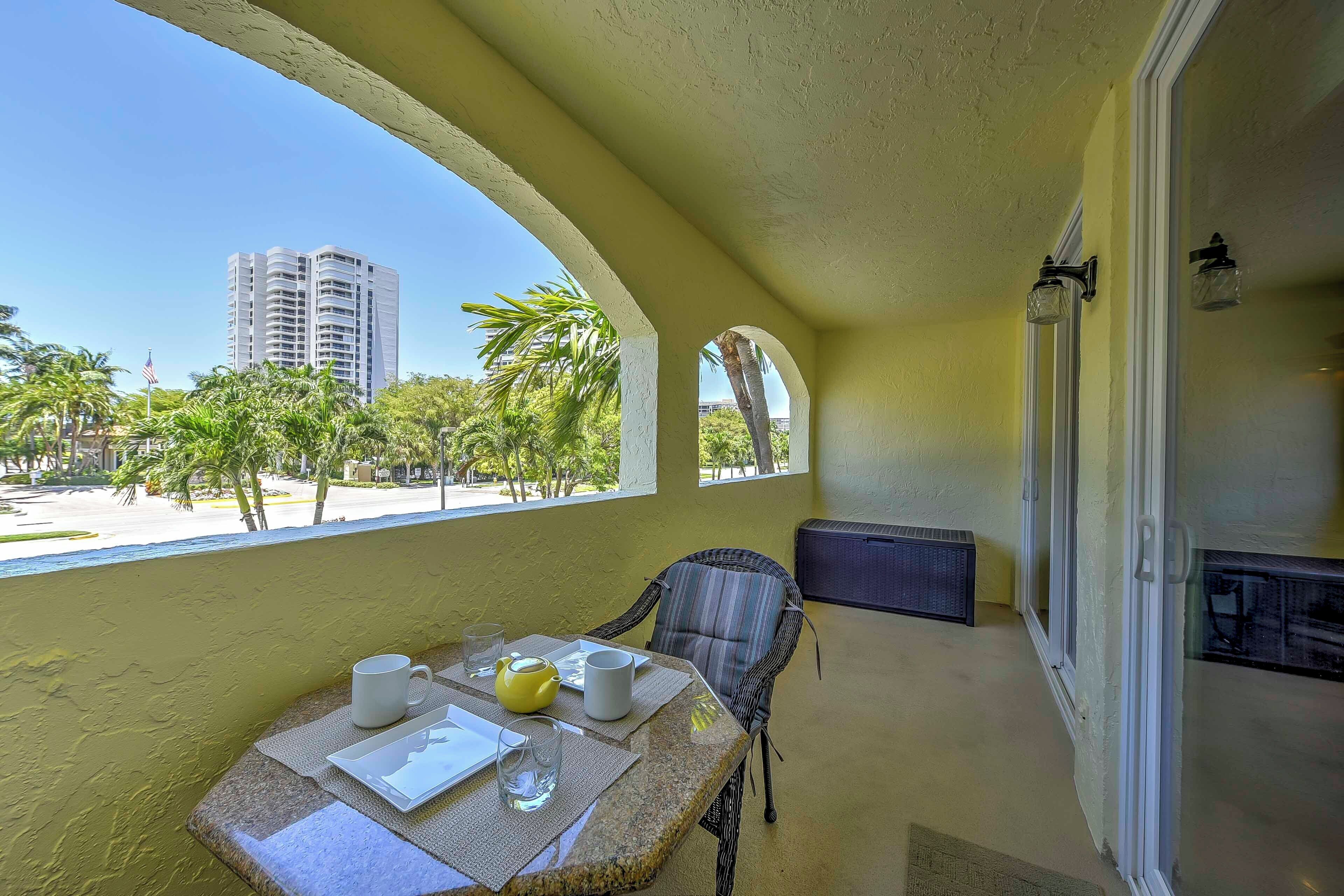 A balcony with a table set for two, overlooking a beach and palm trees.