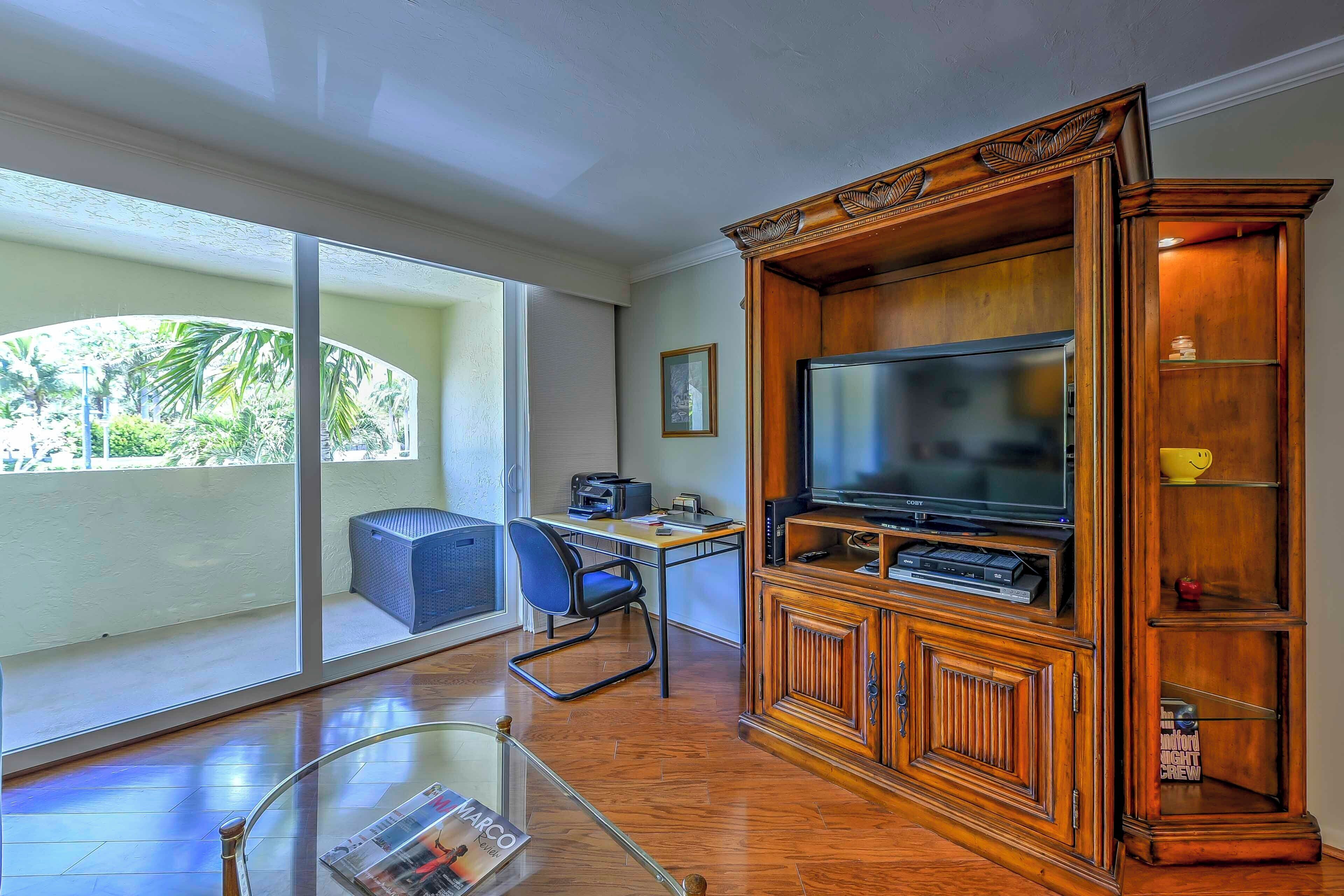 A spacious living room with a large TV, wooden entertainment center, and a view of a balcony with a chair and table.