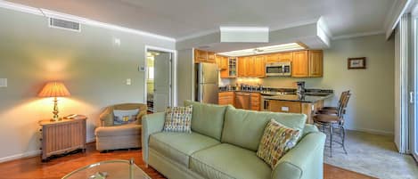 Una sala de estar con un sofá verde, una mesa de centro de cristal y una lámpara. Se ve una cocina al fondo con armarios de madera y un microondas.
