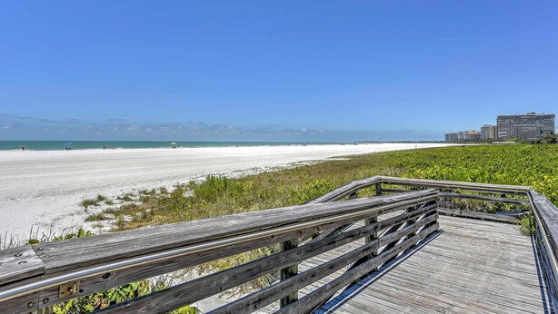 A wooden boardwalk leading to a beach with a clear view of the ocean and distant buildings. - Marco Island Condo w/ Patio: Steps to Beach Access (Marco Island)