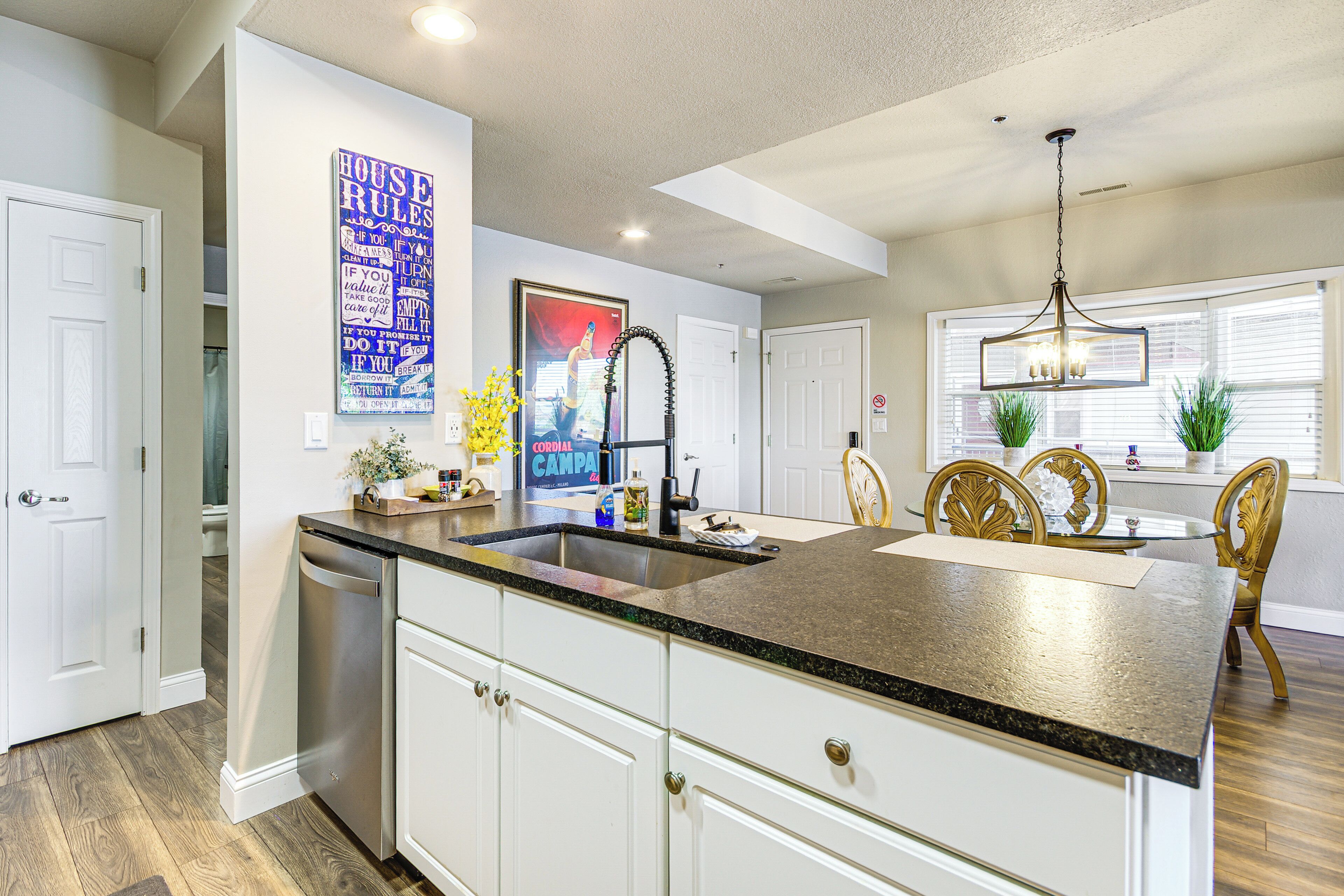 A kitchen with white cabinets, a granite countertop, a stainless steel dishwasher, and a dining area with a glass table and wooden chairs.