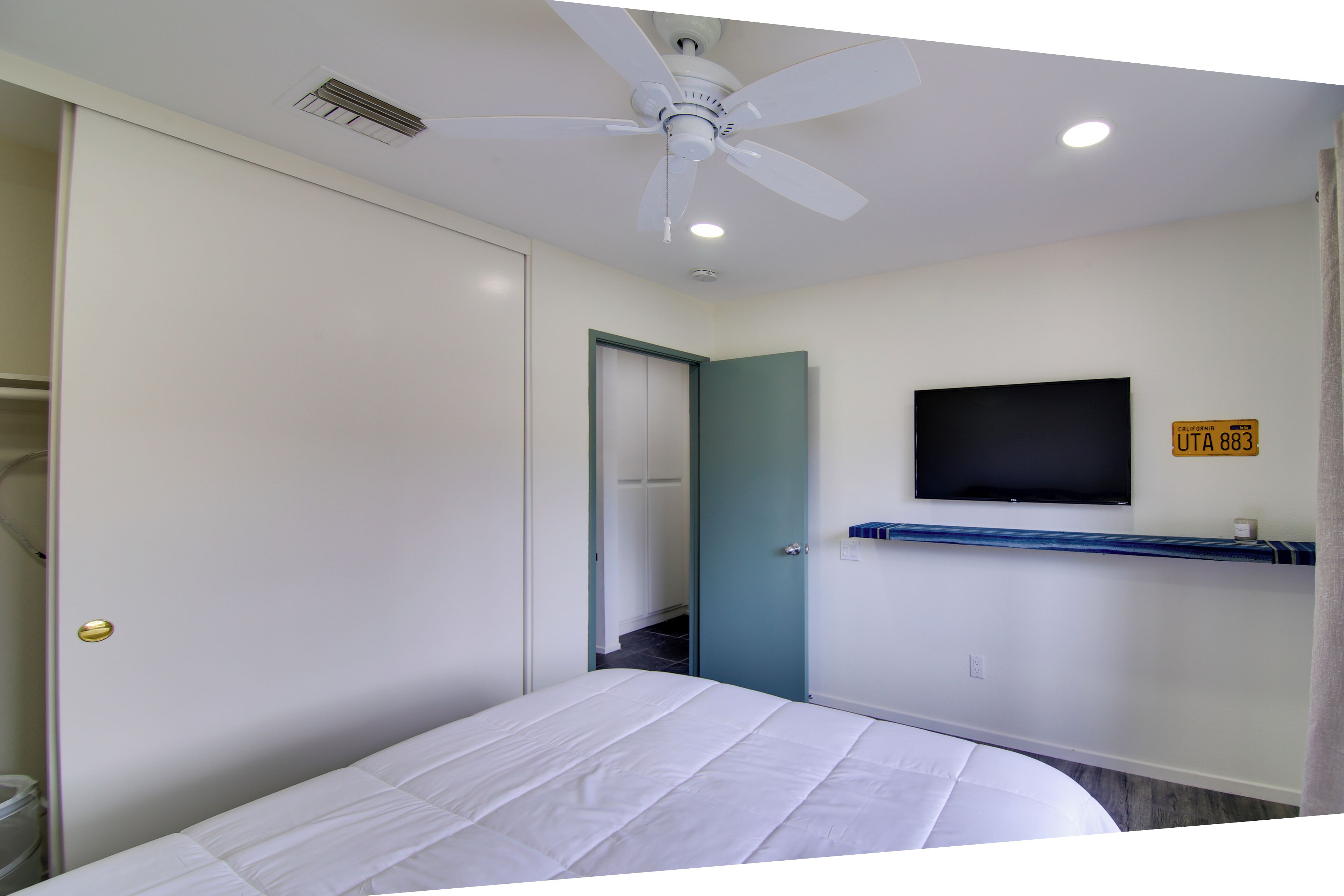 Een slaapkamer met een bed, een plafondventilator, een aan de muur bevestigde televisie en een deur naar een andere kamer.
