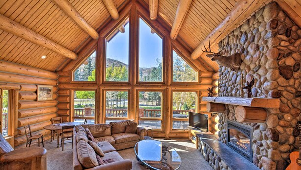 Interior de una cabaña de madera con chimenea de piedra, ventanas grandes, un sofá y un televisor.