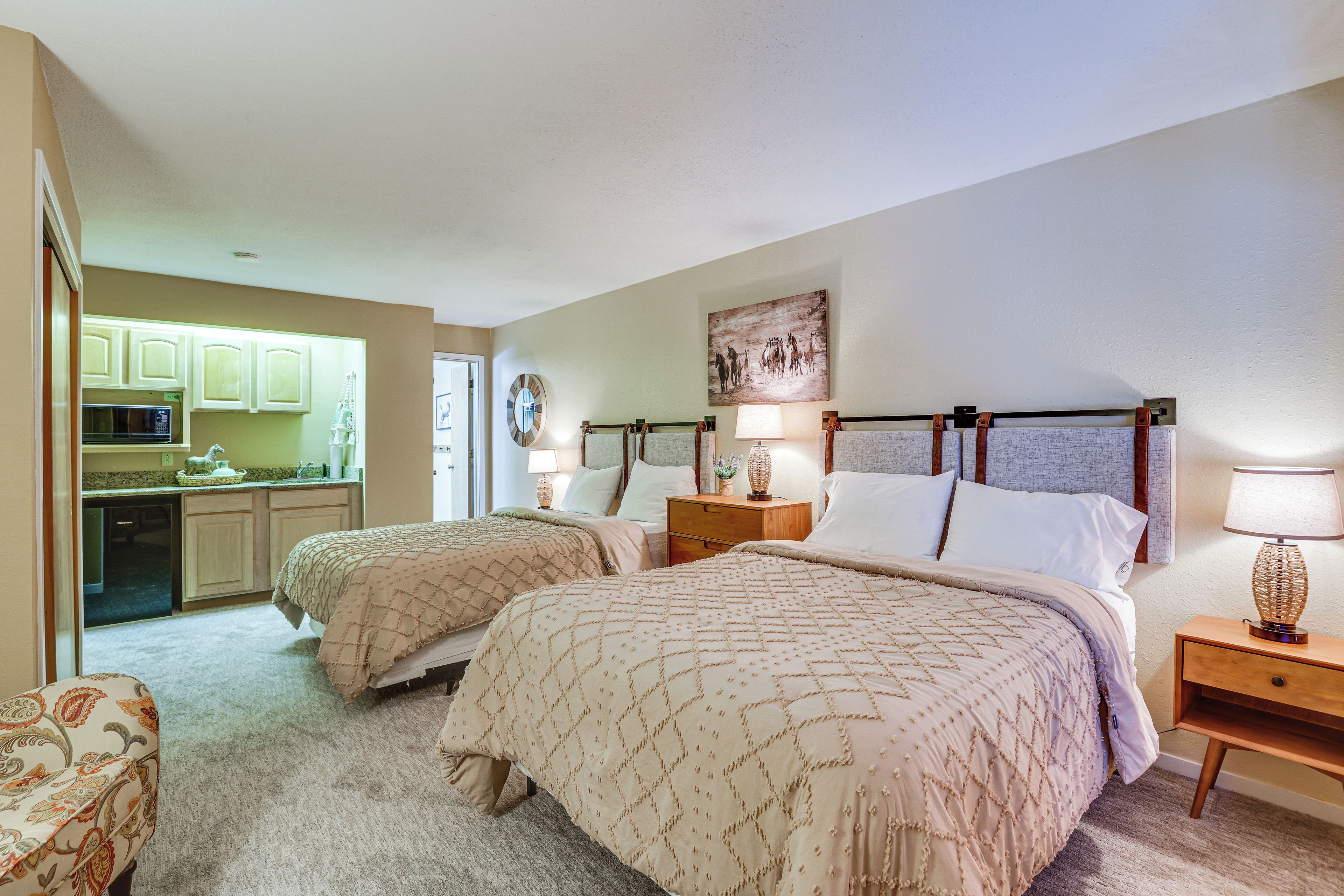 A bedroom with two beds, a nightstand with a lamp, a small table, and a view of a kitchen area.