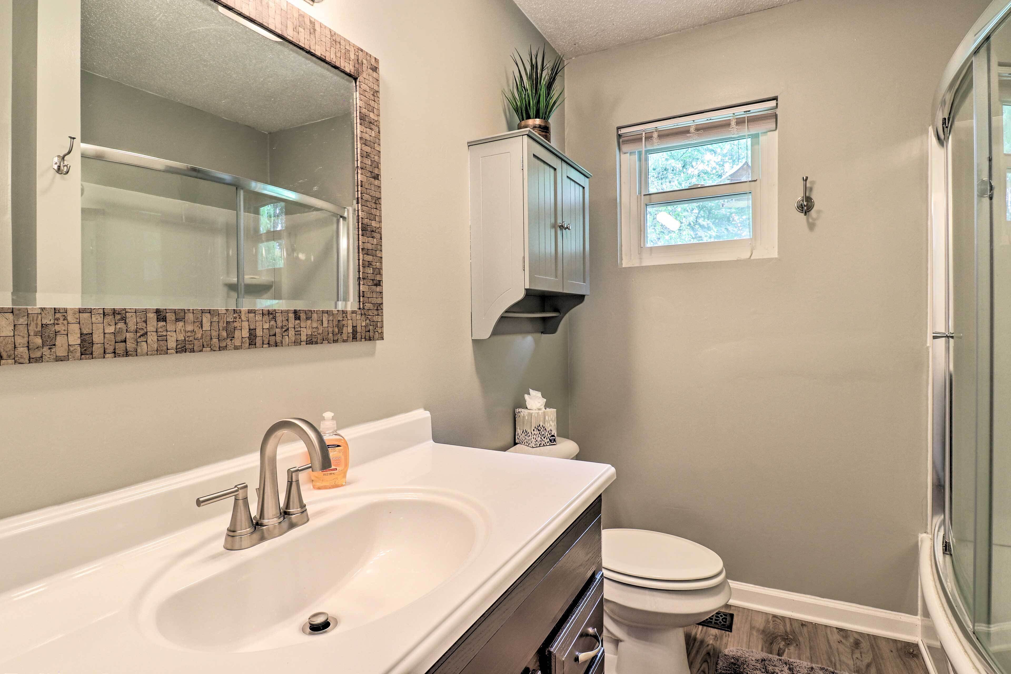 A bathroom with a white sink, a toilet, a glass shower enclosure, a window, and a cabinet with a plant.