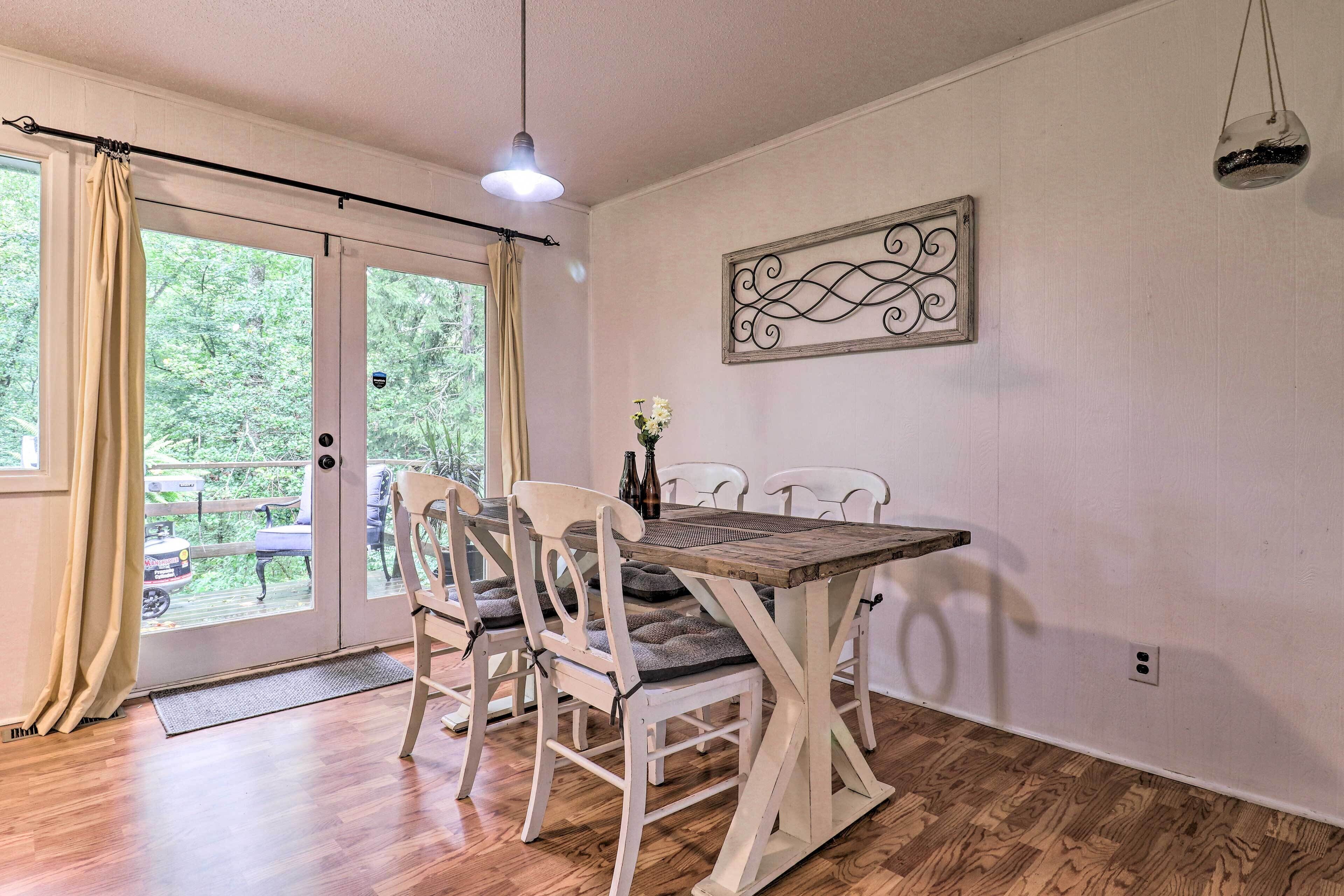 A dining area with a wooden table and white chairs, a decorative wall piece, and a view through glass doors to an outdoor deck.