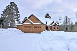 Situated in a wooded area, this two-story house features a central entrance and garage doors, set amidst a serene winter landscape.