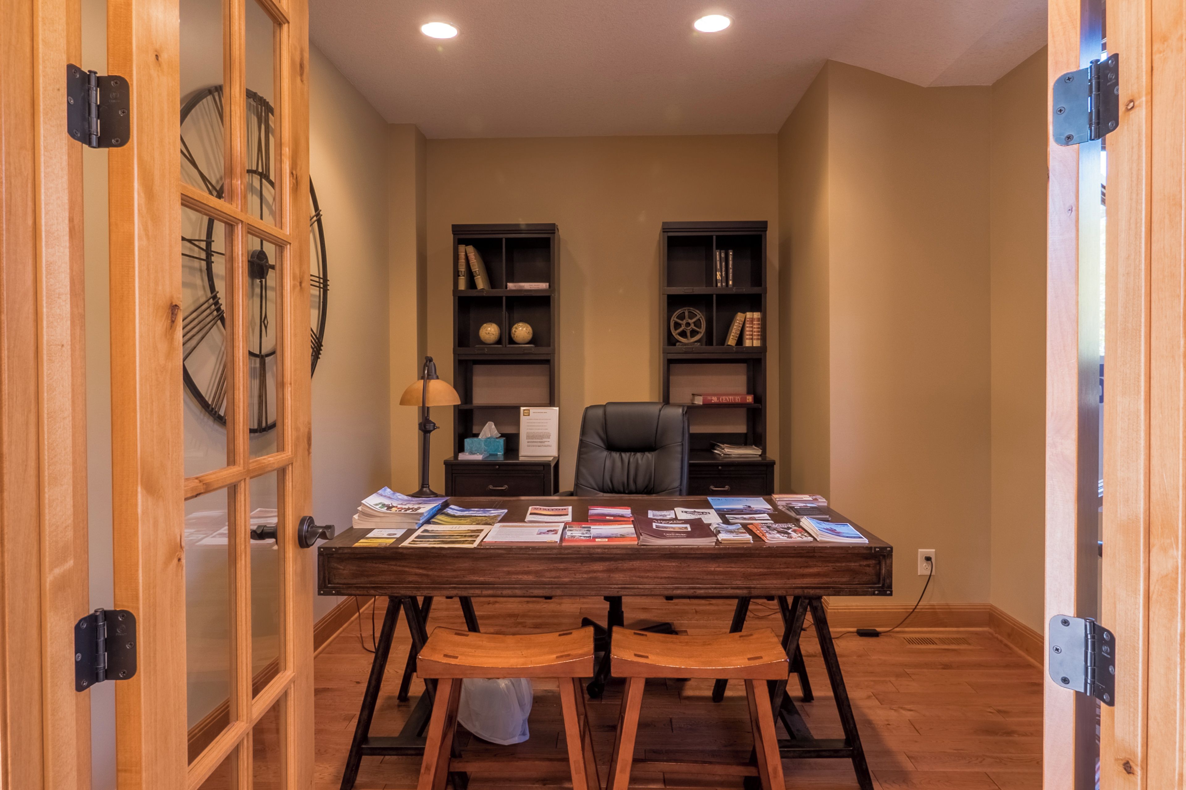 A room with a wooden desk, two chairs, a bookshelf, and a clock on the wall.