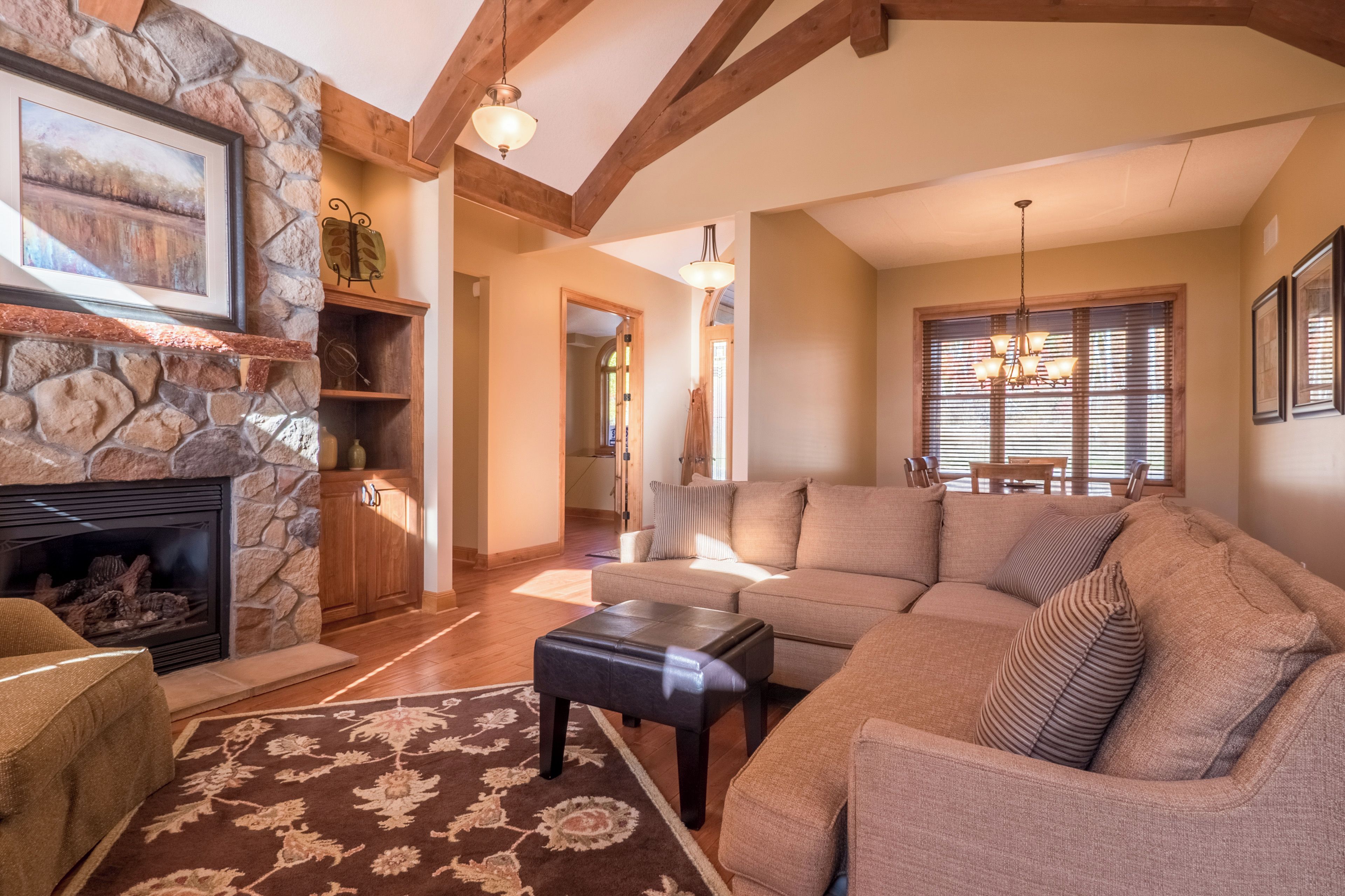 A spacious living room with a stone fireplace, wooden beams, a large sofa, and a dining area with a chandelier.