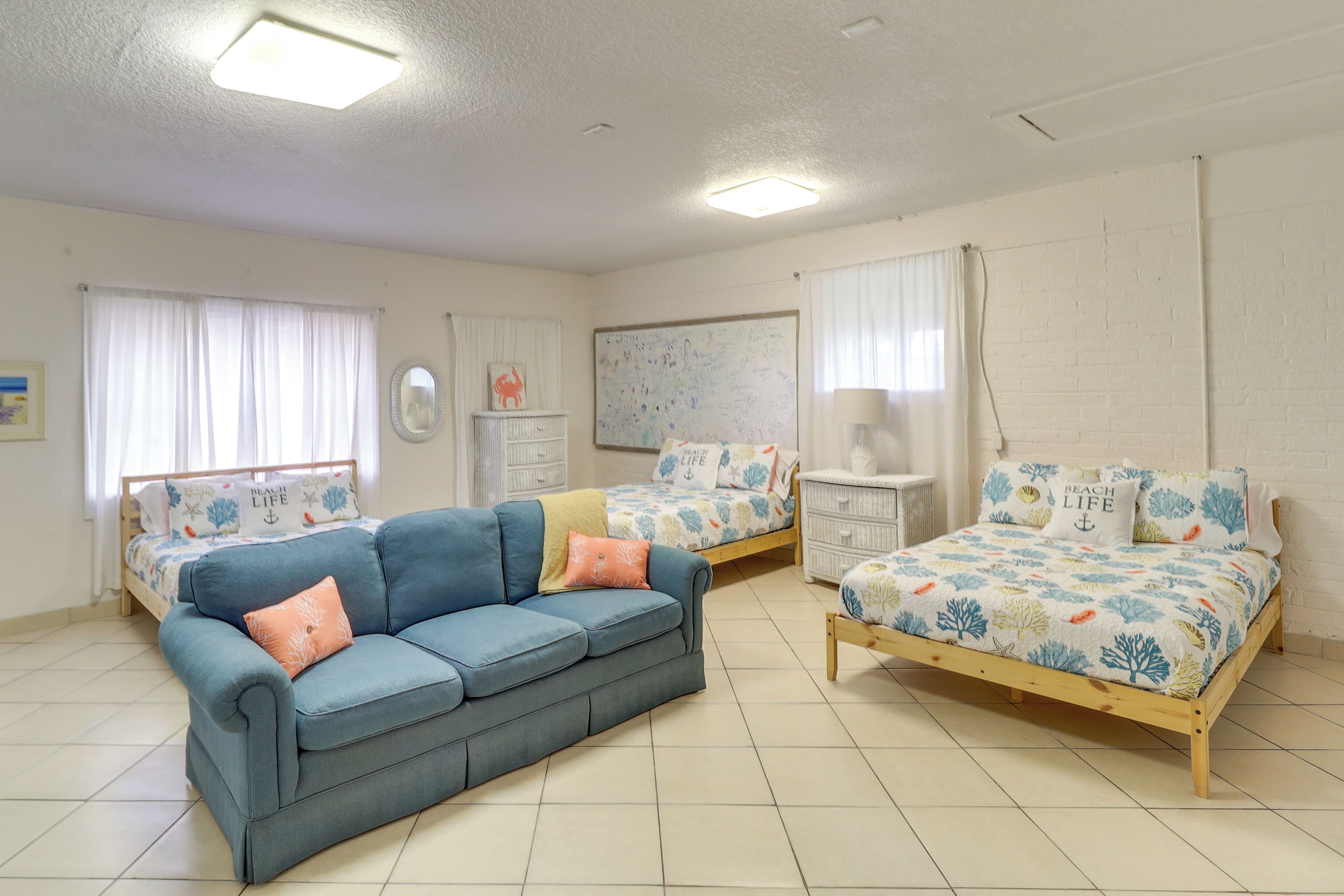Een slaapkamer met een blauwe bank, twee eenpersoonsbedden met bloemenbeddengoed, een witte dressoir en een groot raam met dunne gordijnen.