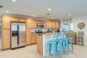A modern kitchen with wooden cabinets, a stainless steel refrigerator, a microwave, and a coffee maker. There are three blue bar stools at the island.