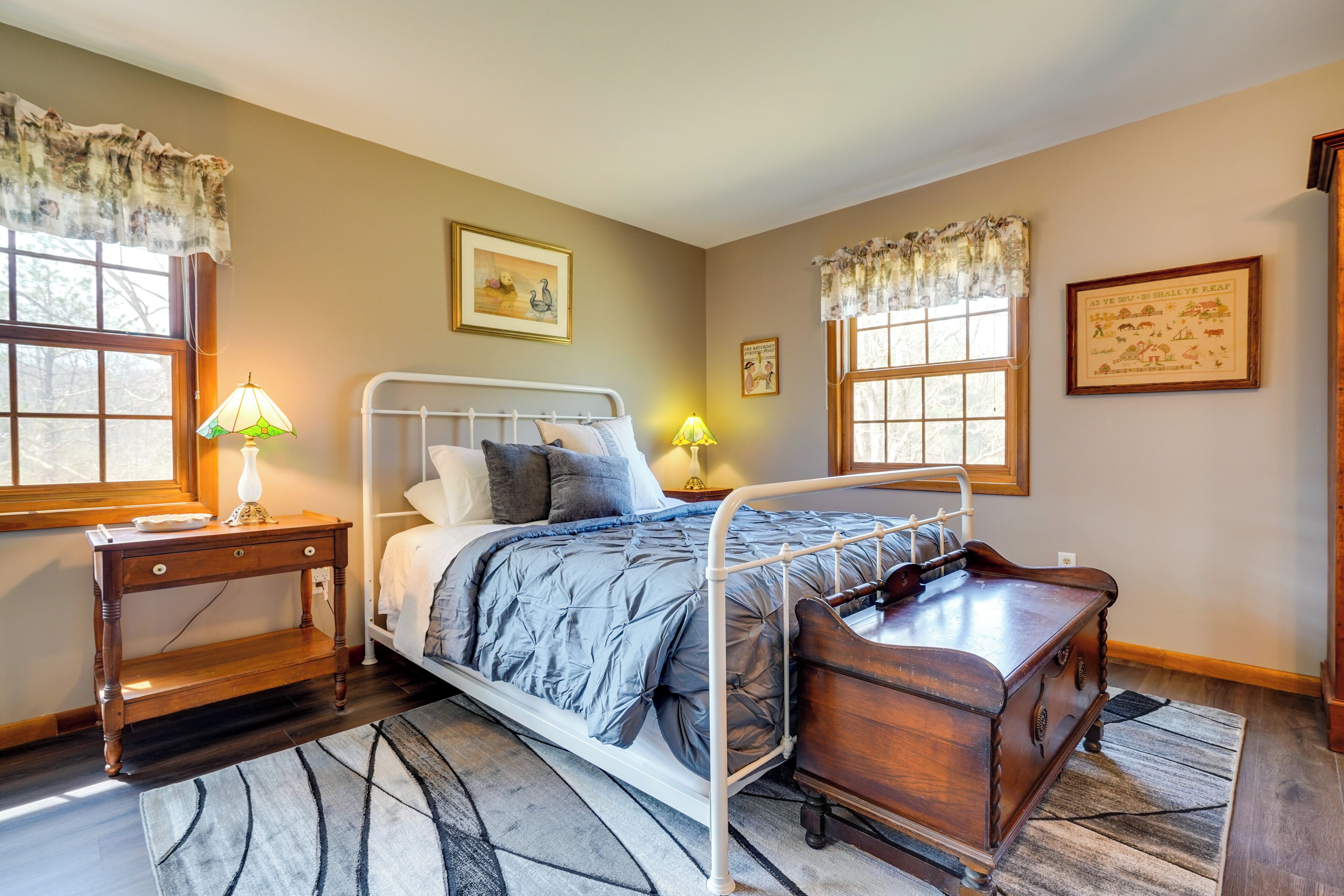 A bedroom with a metal bed frame, a wooden nightstand, a bedside table, and a dresser. There are two windows with curtains, a rug on the floor, and wall art.
