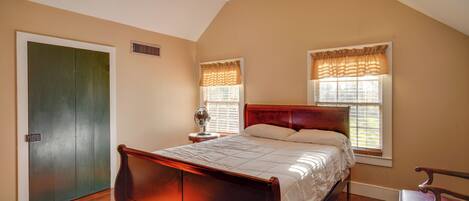 A bedroom with a wooden bed, two windows with curtains, a bedside table, and a chair.