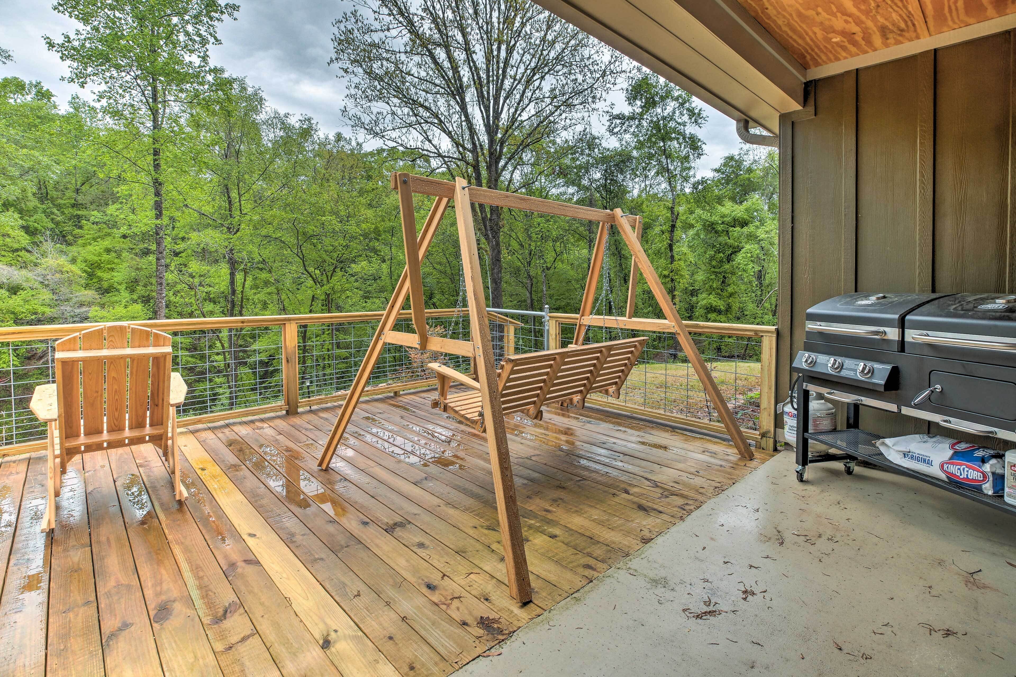 Une terrasse en bois avec une balançoire, un barbecue et un espace abrité offrant une vue sur une forêt.