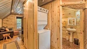 Una cabina compatta con arredamento in legno, lavatrice e asciugatrice, un bagno piccolo con lavabo e specchio, e un angolo salotto accogliente con divano e poggiapiedi.