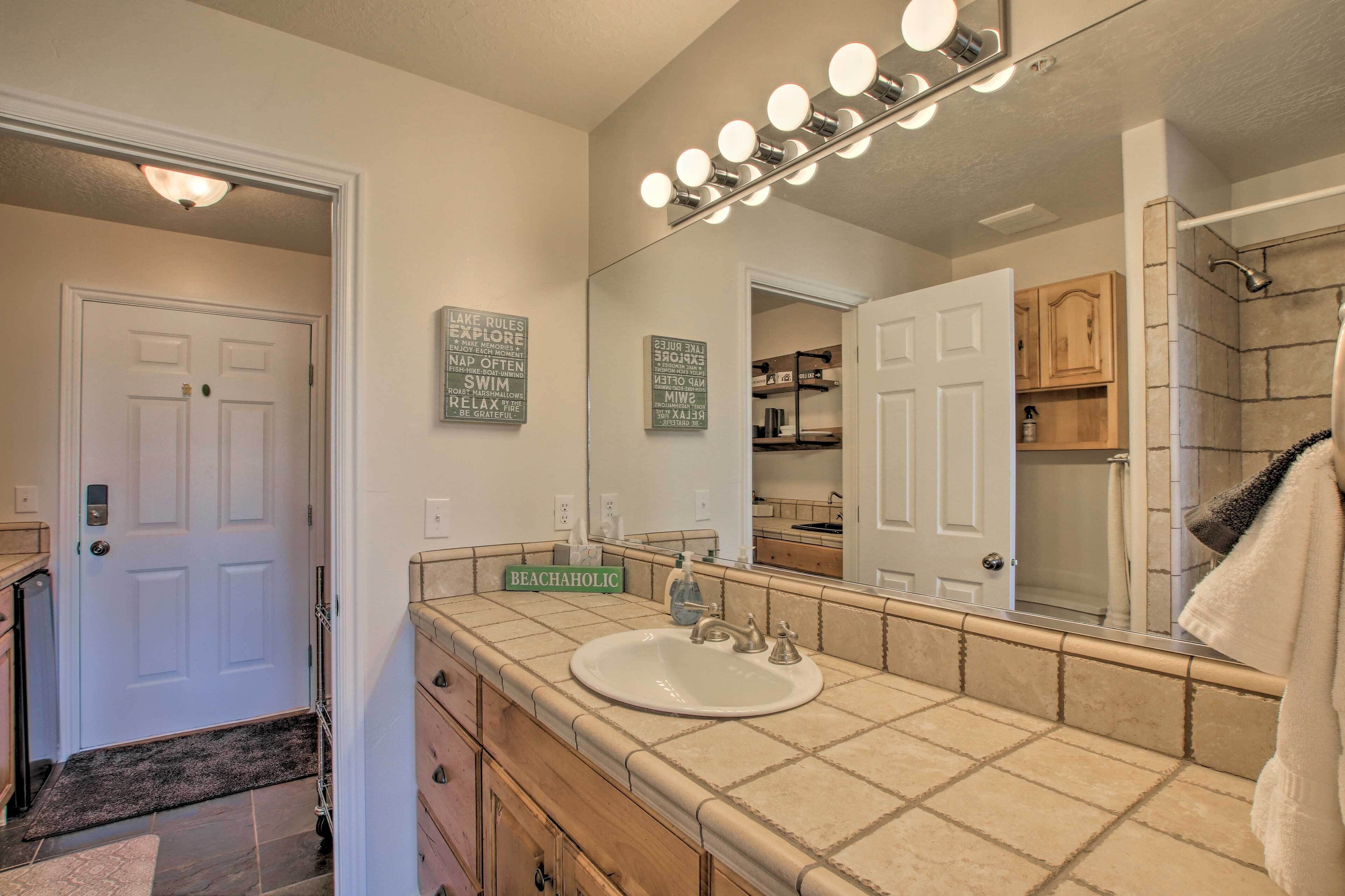 A bathroom with a white sink, vanity, and a large mirror with lights. There are motivational signs on the wall and a towel rack.