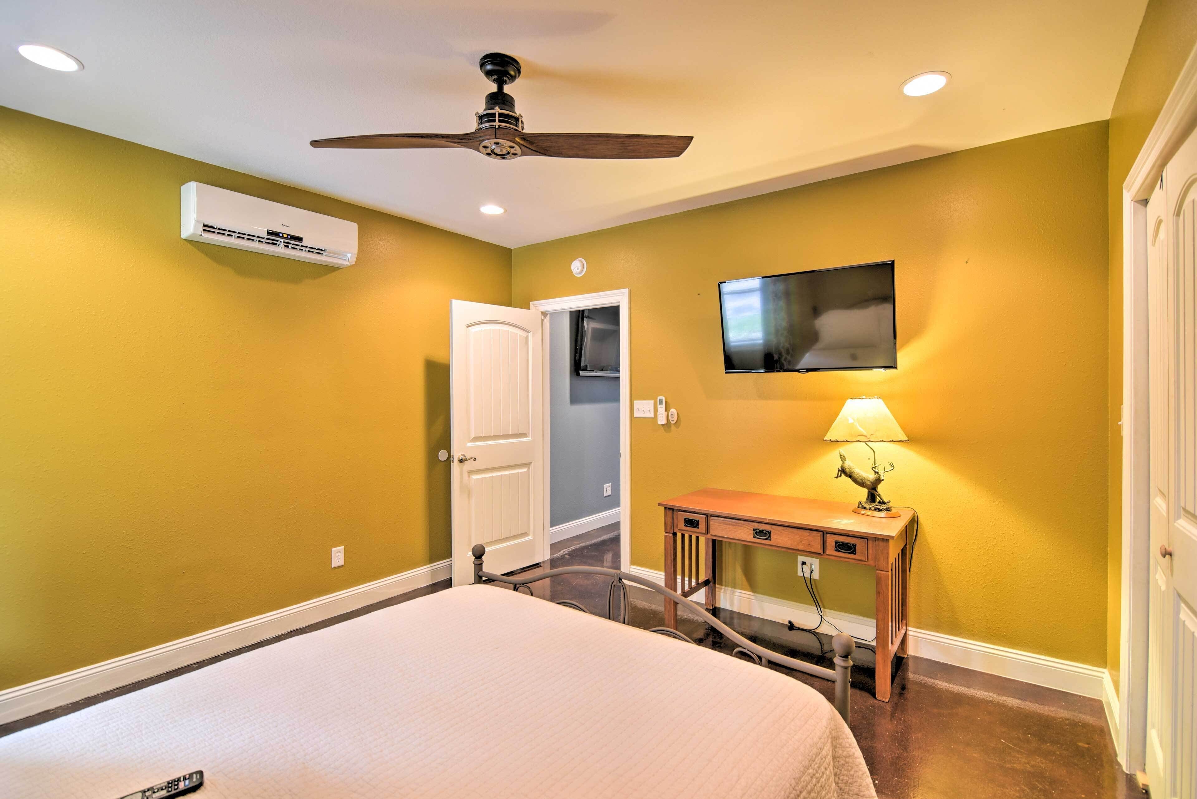 A hotel room with a bed, ceiling fan, air conditioning unit, television, bedside table with lamp, and a door to the bathroom.