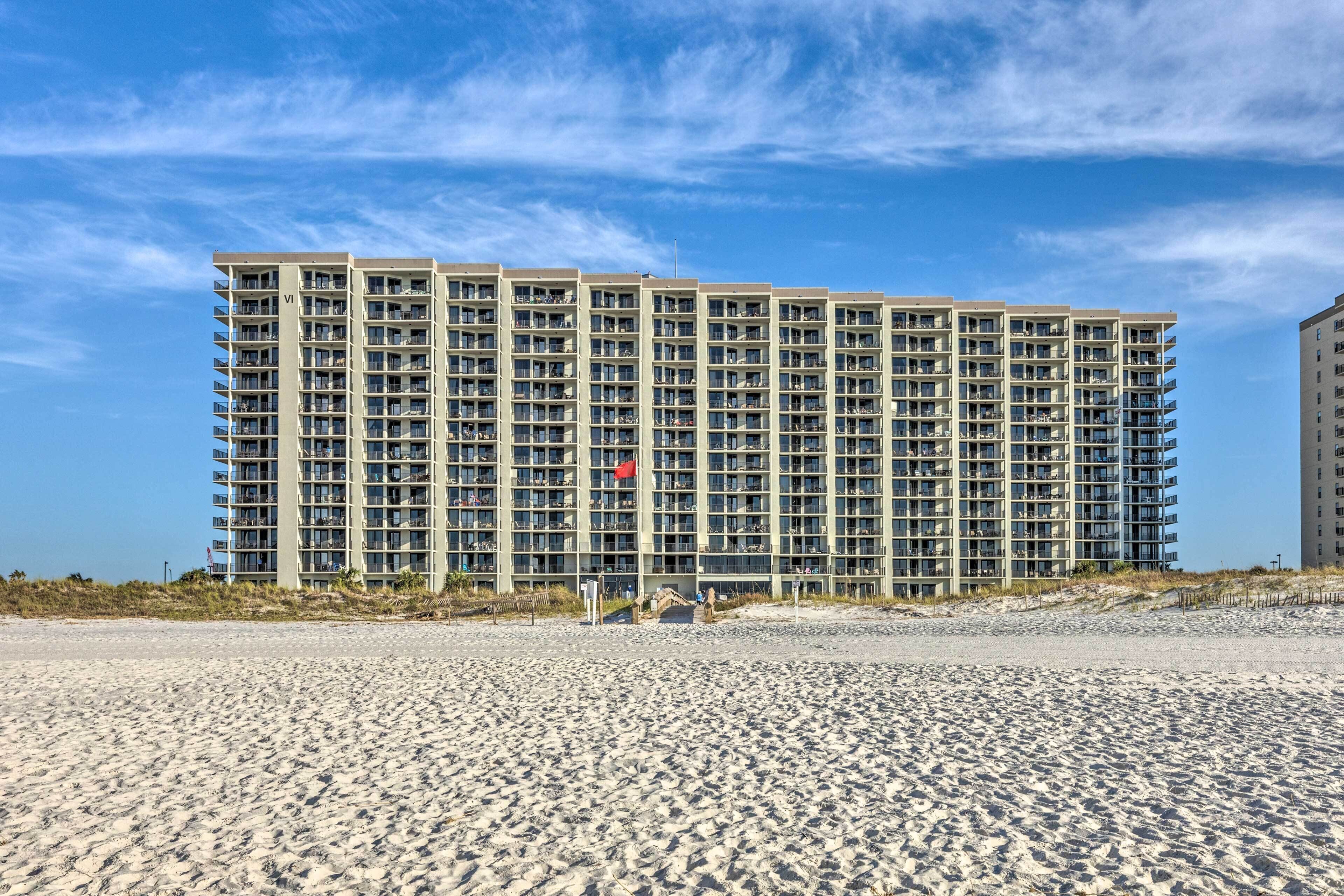 A high-rise hotel with multiple floors, a beach in the foreground, and a clear sky.