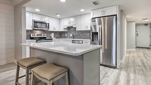 A modern kitchen with white cabinets, stainless steel appliances, and a central island with stools. - Coastal Orange Beach Condo: Resort + Beach Access! (Orange Beach)