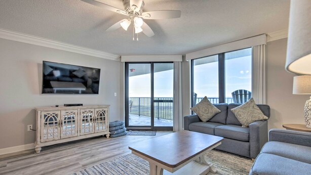 A living room with a ceiling fan, a flat-screen TV, a grey sofa with cushions, a wooden coffee table, and a large window with a view of a balcony. - Coastal Orange Beach Condo: Resort + Beach Access! (Orange Beach)