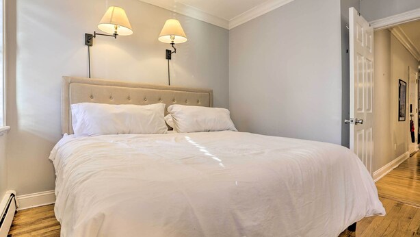A neatly made bed with white linens, two wall-mounted lamps, and a wooden floor. - Couples Hub Near Metlife, Walk to Public Transit! (Hoboken)