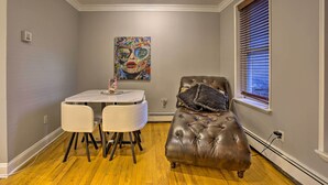 A dining area with a marble table and two chairs, a tufted leather sofa with a throw pillow, a painting on the wall, and a window with blinds. - Couples Hub Near Metlife, Walk to Public Transit! (Hoboken)
