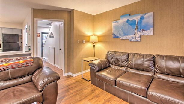 A living room with a brown leather sofa, a small side table with a lamp, and a wall-mounted ski-themed artwork. - 2 Blocks to Skiing & Mtb: Angel Fire Condo (Angel Fire)