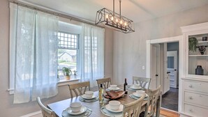 A dining room with a wooden table set for six, white curtains, a chandelier, and a view of the outside.