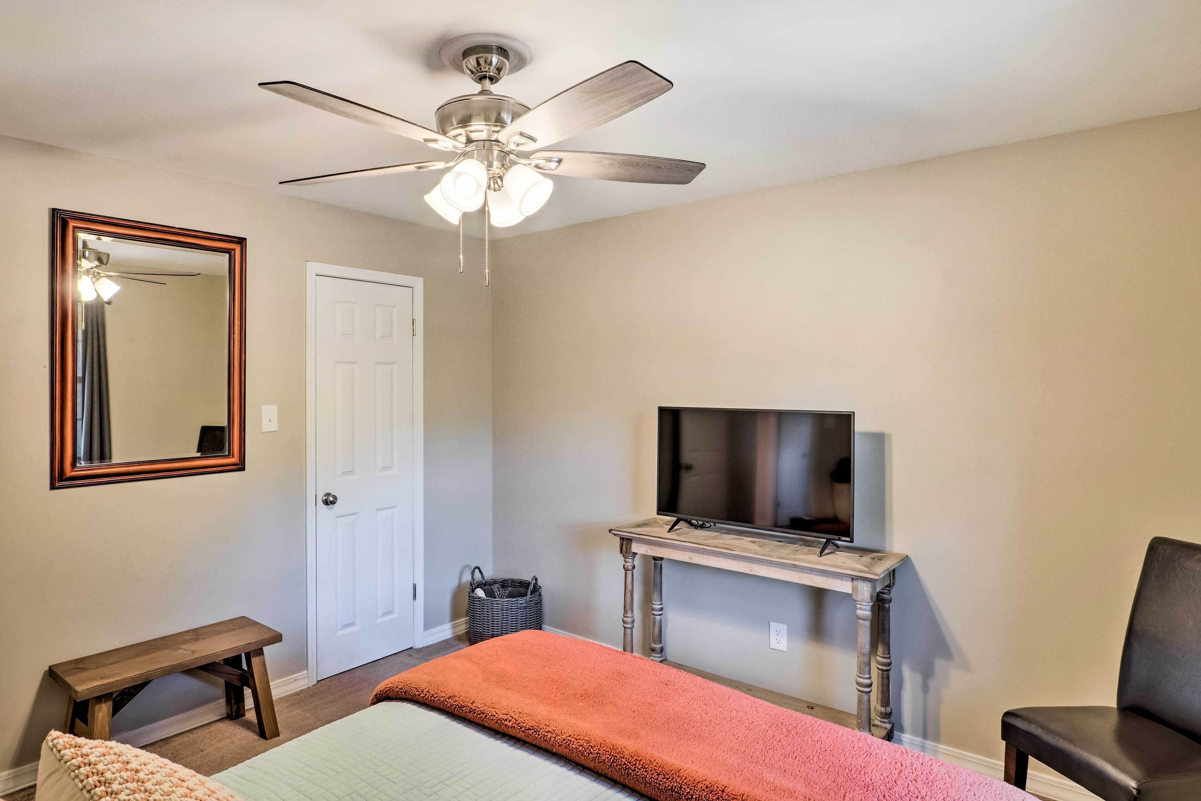 A bedroom with a bed, a ceiling fan, a television on a wooden stand, a bench, and a door.