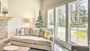 A living room with a beige sofa, a fireplace, a lamp, and a view of a wooded area through large windows.