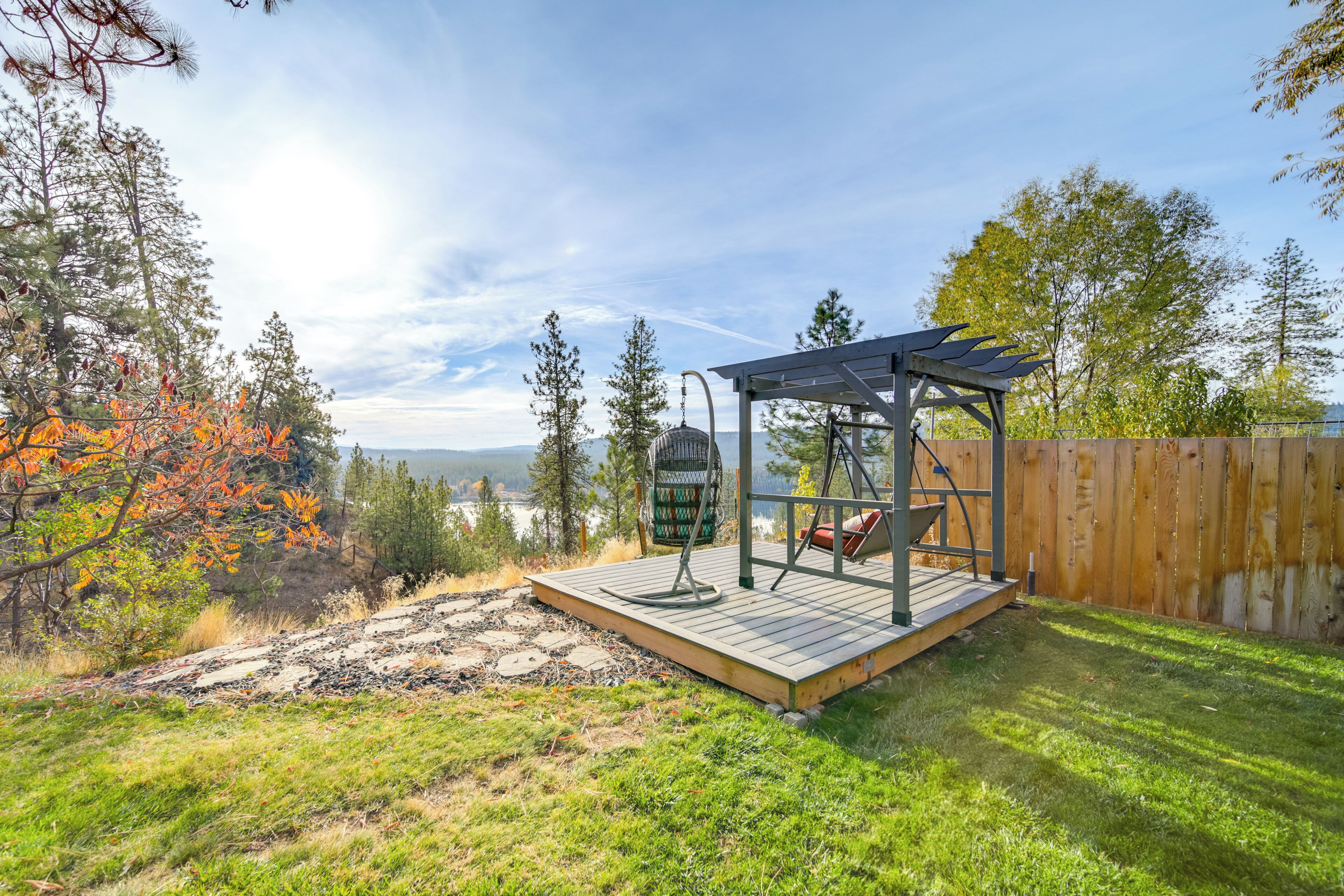 A wooden deck with a swing set and a pergola, surrounded by trees and a view of a lake.