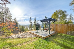 Une terrasse en bois avec une balançoire et une pergola, entourée d’arbres et offrant une vue sur un lac.