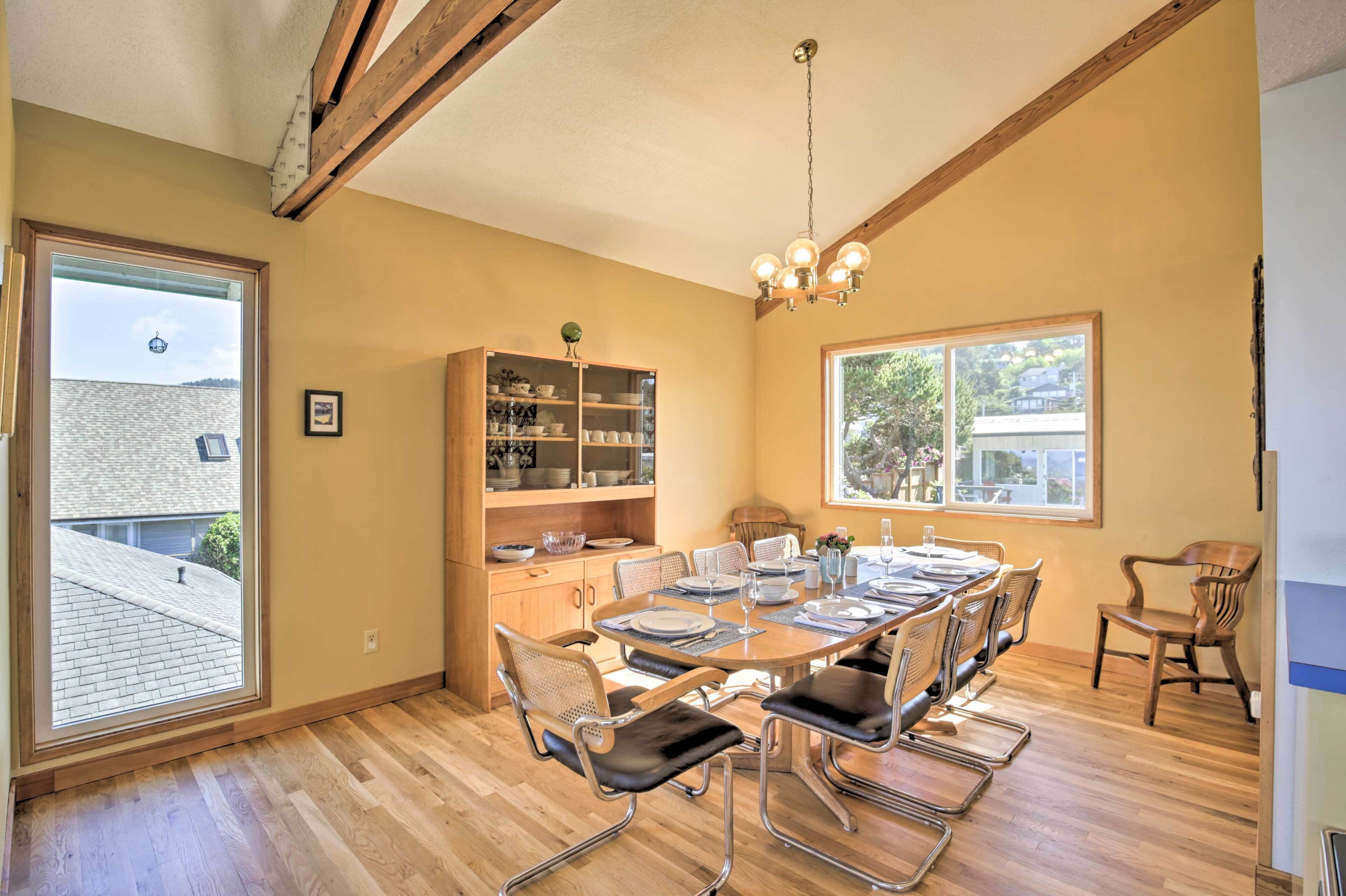 A dining area with a wooden table set for six, surrounded by chairs with metal frames and leather seats. A wooden cabinet with glass doors is against the wall, and a chandelier hangs from the ceiling.