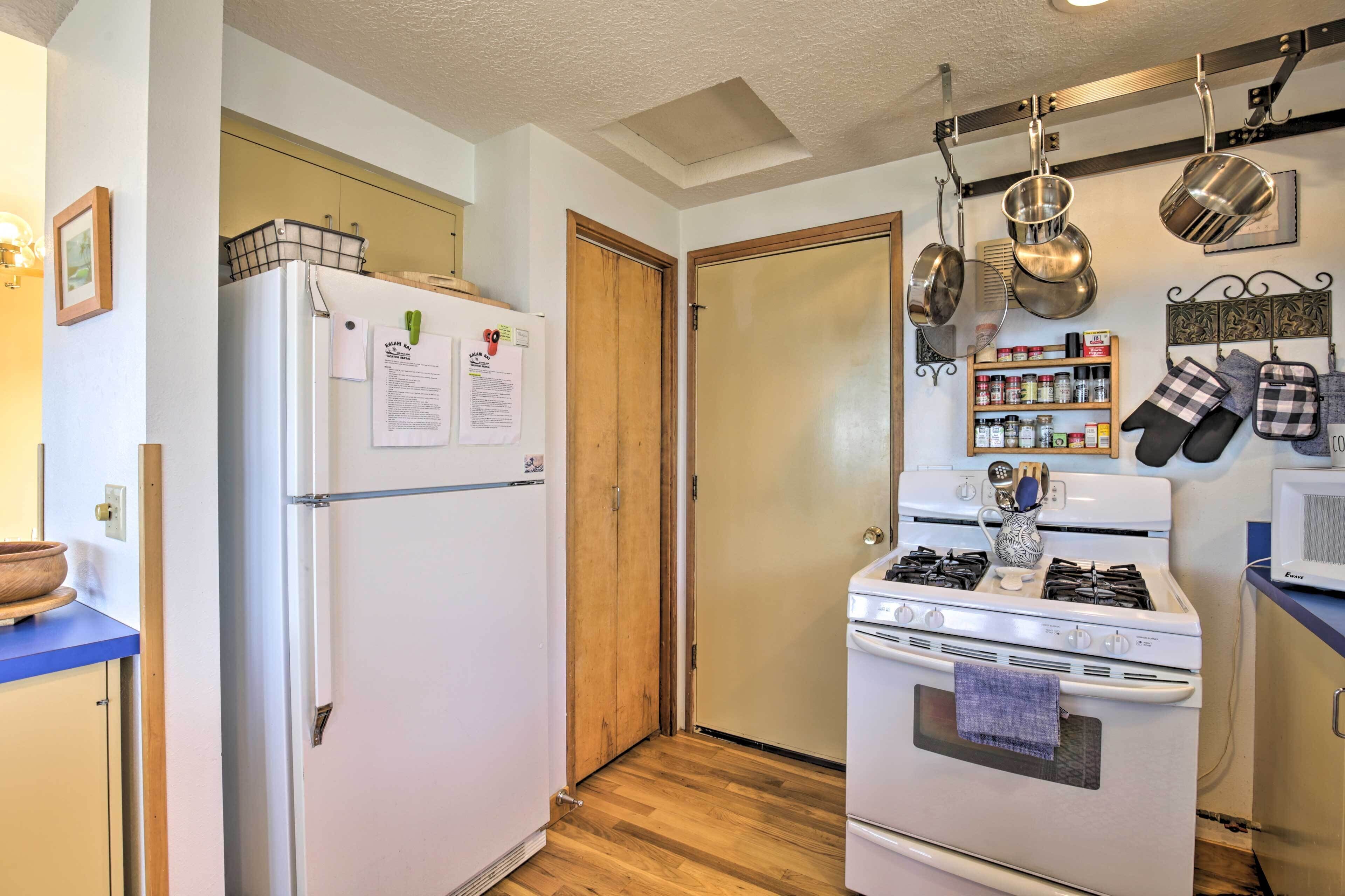 A compact kitchen with a refrigerator, stove, and microwave. There are hanging pots and a spice rack.
