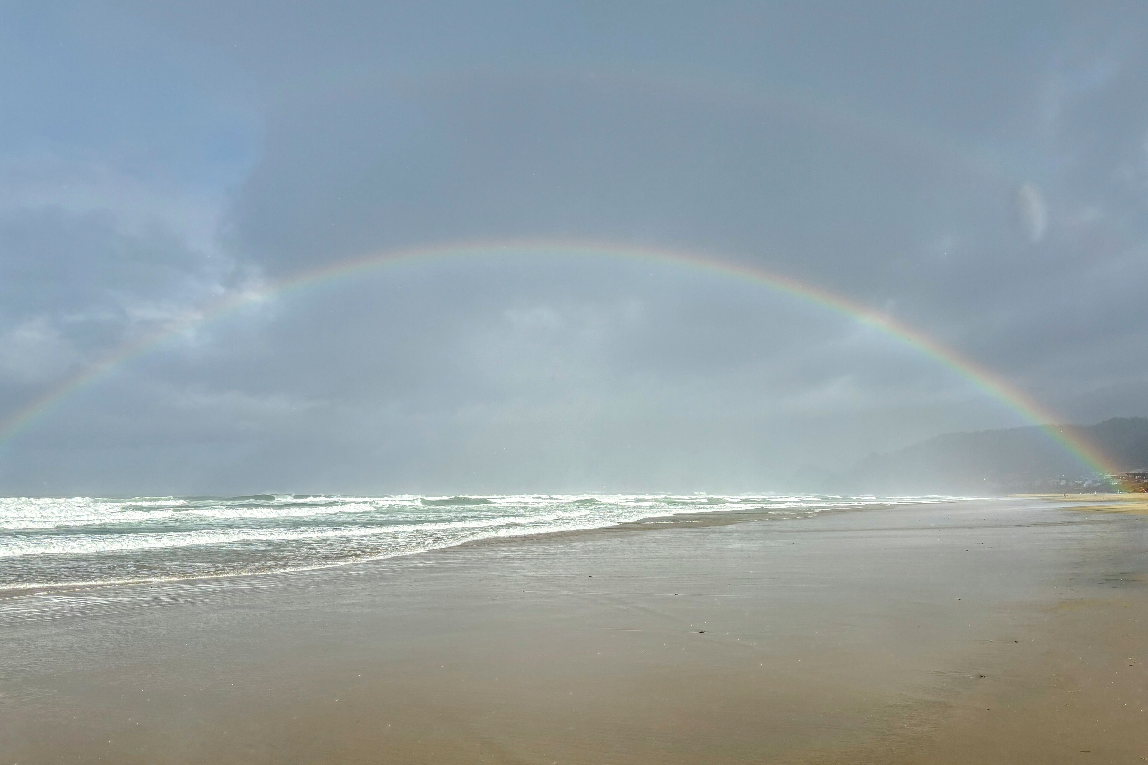 A beach with a rainbow over the ocean and mountains in the background.