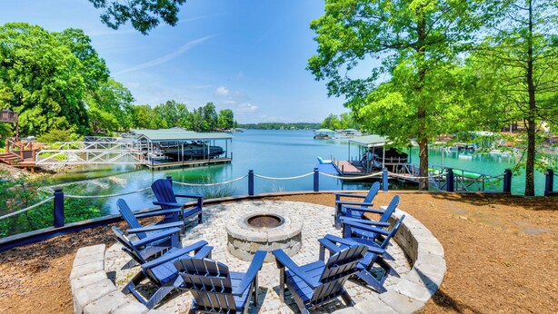 A lakeside patio with Adirondack chairs, a fire pit, and a dock extending into the water. - Beautiful Lake Keowee Home w/ Boat Dock & Fire Pit (Seneca)