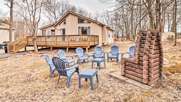 Une maison en bois avec une terrasse, entourée d’arbres. On y trouve plusieurs chaises et un foyer.