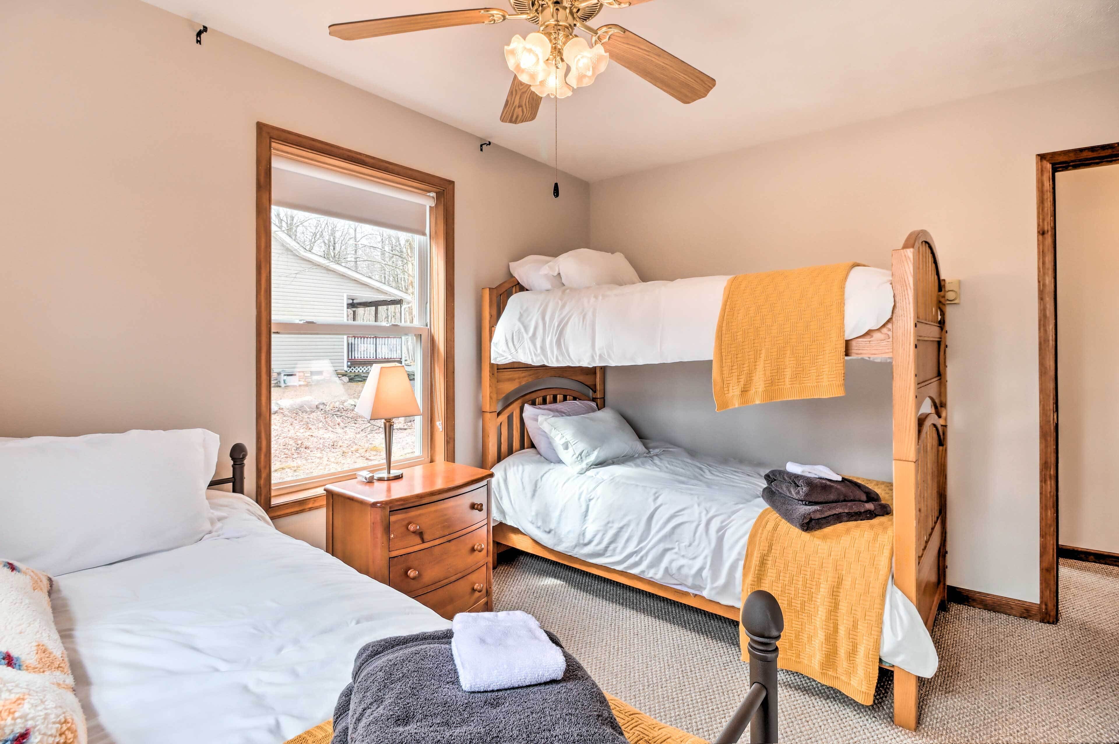 A bedroom with a bunk bed, a single bed, a wooden nightstand, a lamp, and a window with a view of a house.