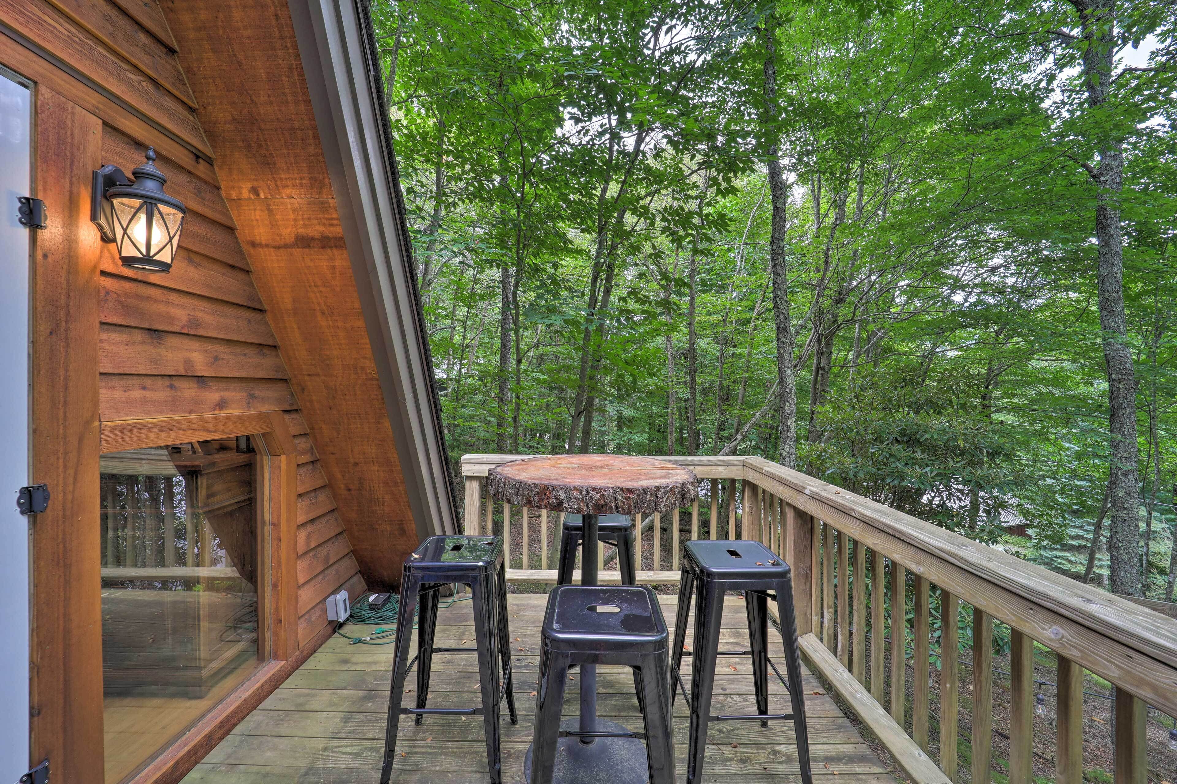 A wooden deck with a table and stools, overlooking a forested area.