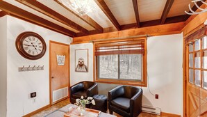 A cozy room with a wooden ceiling, a large window with blinds, two black leather chairs, a wooden table with a tray, a wall clock, and a framed picture. - Private Hot Tub: Cabin 1 Mi to Beech Mountain! (Banner Elk)