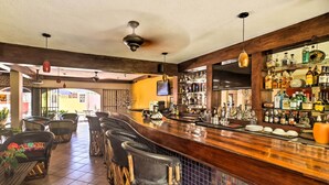 Una zona de bar con sillas de madera, suelo de baldosas, barra con botellas y una pantalla de televisión.